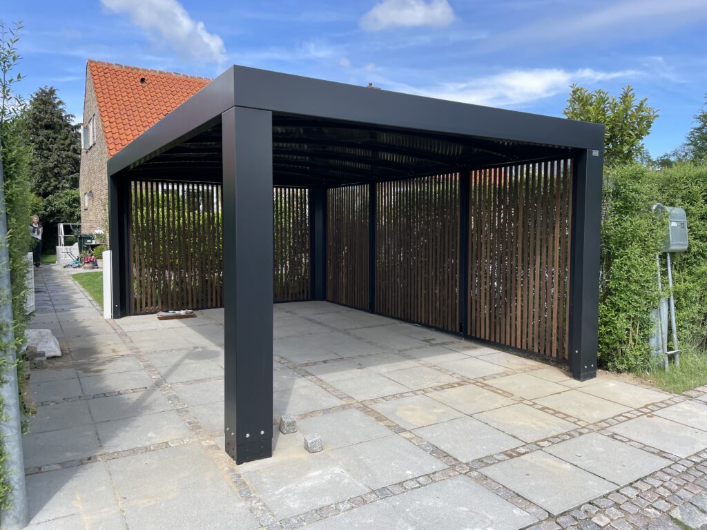 Sort stålcarport fra Allview Carporte med lamelvægge, monteret på flisebelagt grund ved et moderne hus.