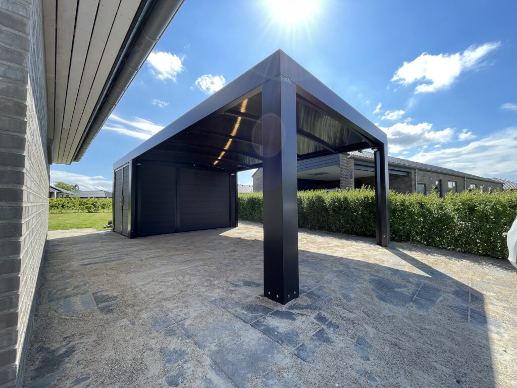 Sort stålcarport med endevæg fra Allview Carporte, monteret ved et hus med flisebelagt grund og hæk.